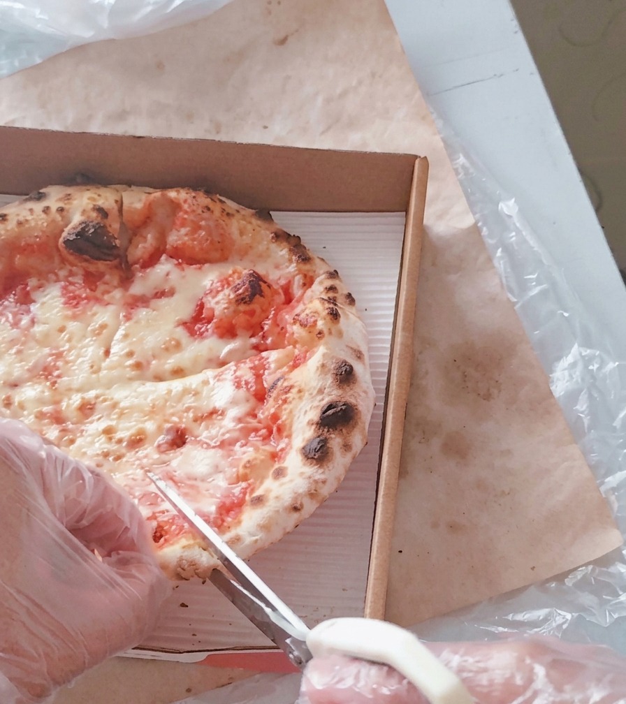 Una persona con guantes de plástico usando unas tijeras de cocina blancas para cortar una pizza de queso caliente en porciones dentro de una caja de cartón.