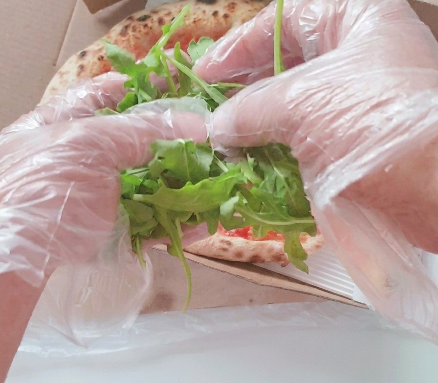 Manos con guantes de plástico transparentes colocando un montón de hojas de rúcula verde fresca en el centro de una pizza horneada dentro de una caja.