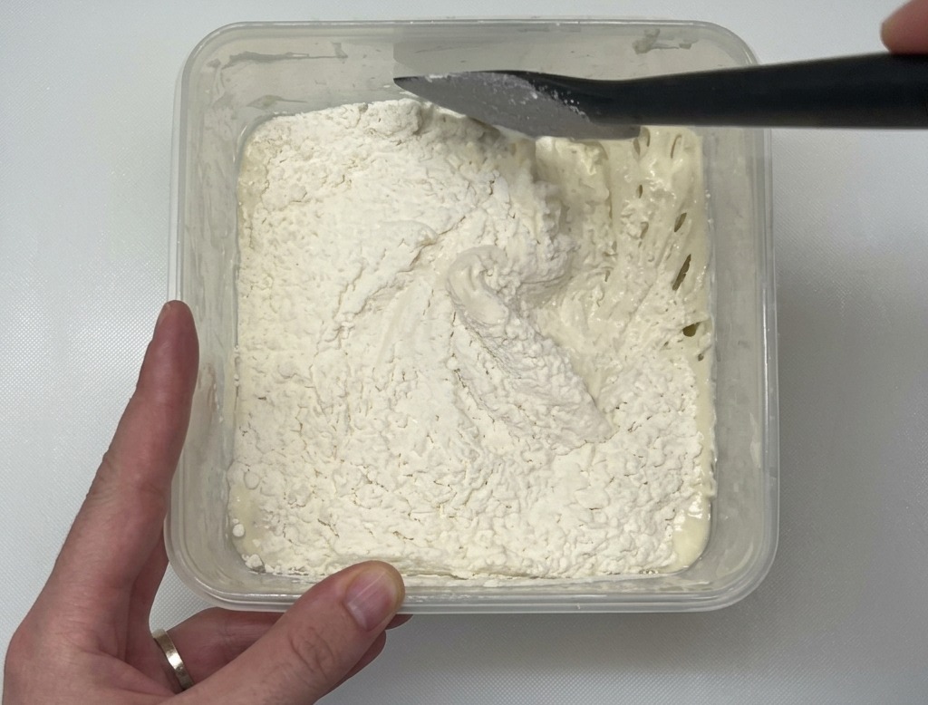 Una espátula negra mezclando harina blanca en una masa húmeda dentro de un recipiente de plástico transparente sobre una encimera blanca.