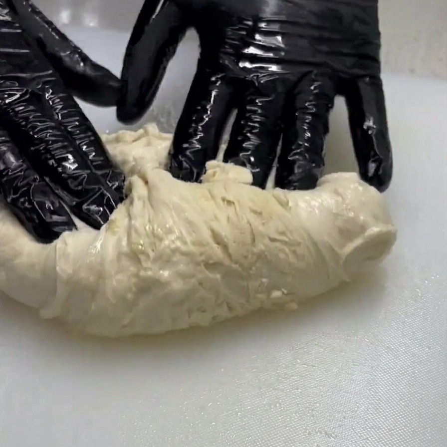 Una persona con guantes negros estirando y plegando una porción de masa pálida y húmeda sobre una superficie blanca.