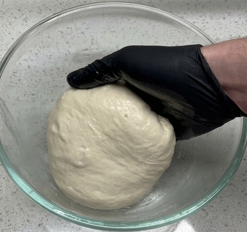 Una mano con guante negro colocando una bola de masa lisa en un bol grande de cristal transparente sobre una encimera de estilo granito.