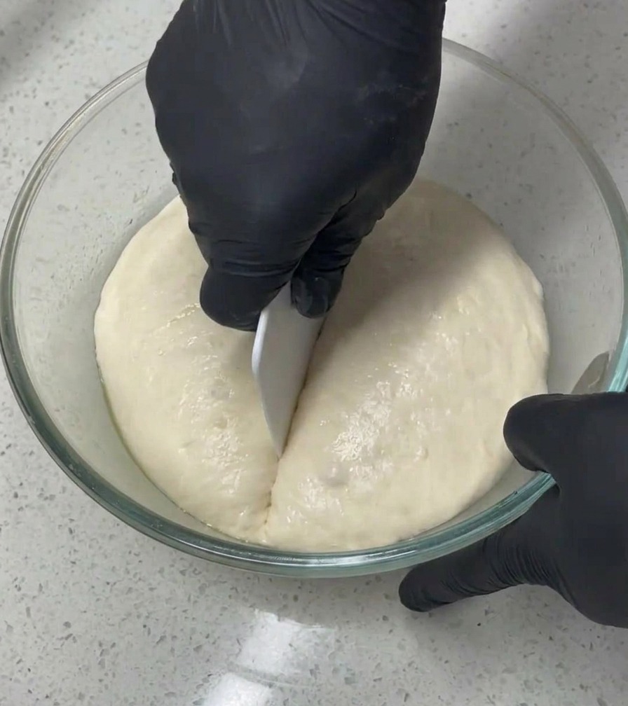 Una persona con guantes negros utiliza una rasqueta blanca para cortar por la mitad una masa grande y aireada dentro de un bol de cristal.