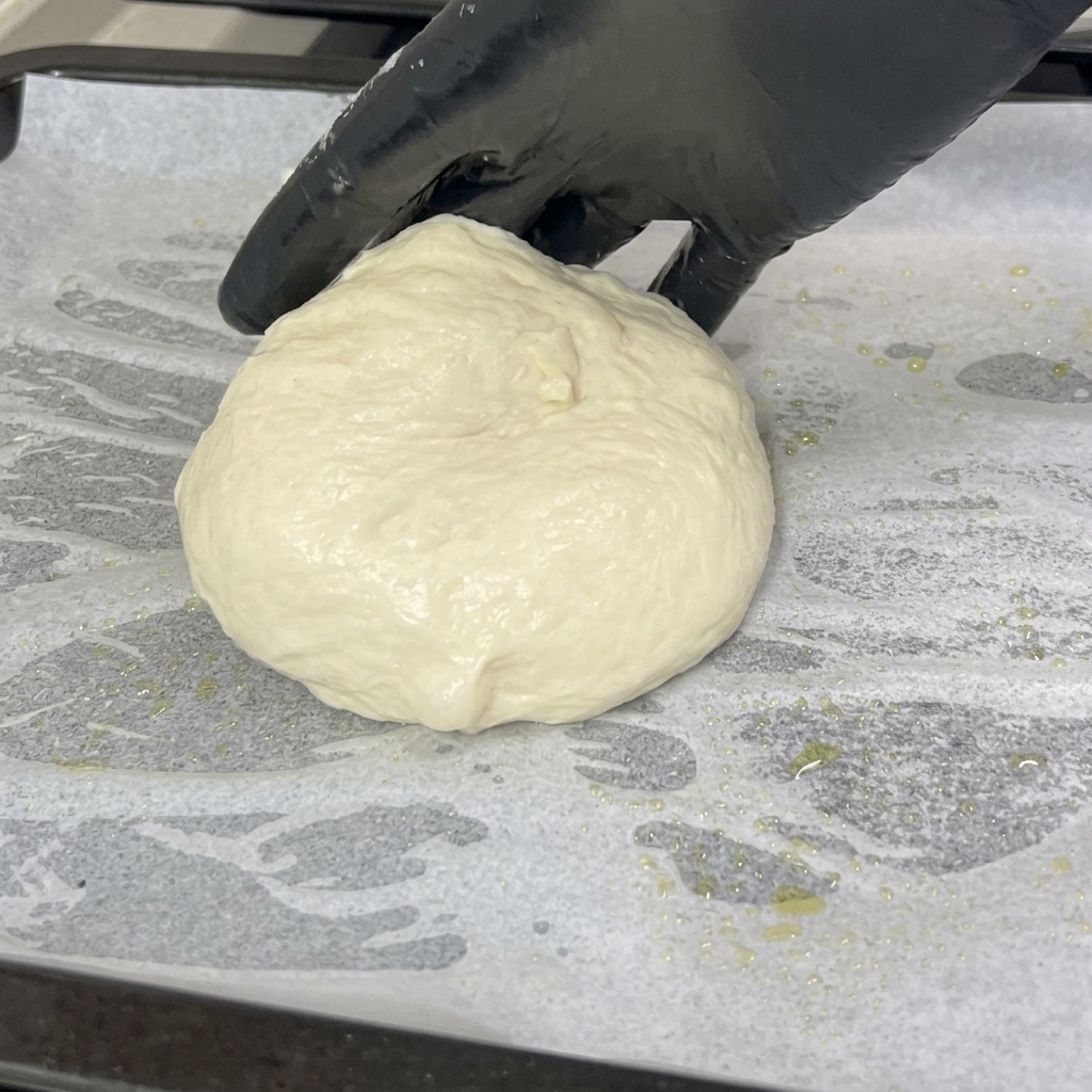 Una mano con guante coloca una bola de masa de pizza suave y redondeada sobre una bandeja cubierta con papel de horno blanco.