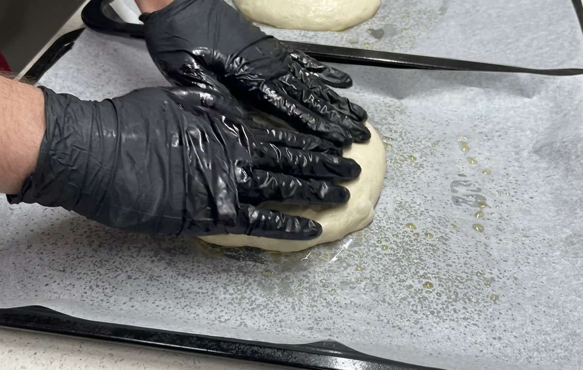 Dos manos con guantes negros presionan sobre una porción de masa extendida en una bandeja de horno para llevarla hacia los bordes.