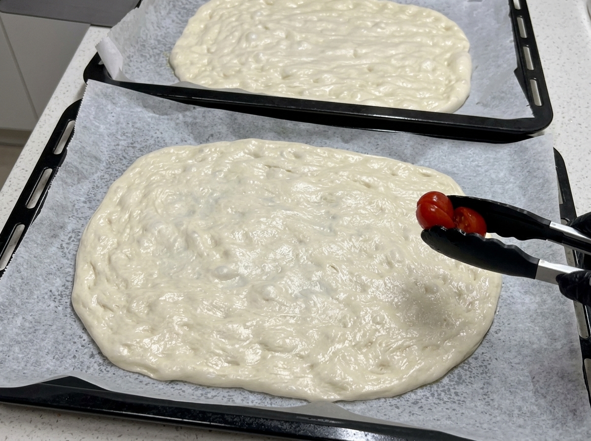 Con unas pinzas de cocina negras, se coloca la mitad de un tomate cherry sobre una base grande de masa de pizza cruda y extendida.