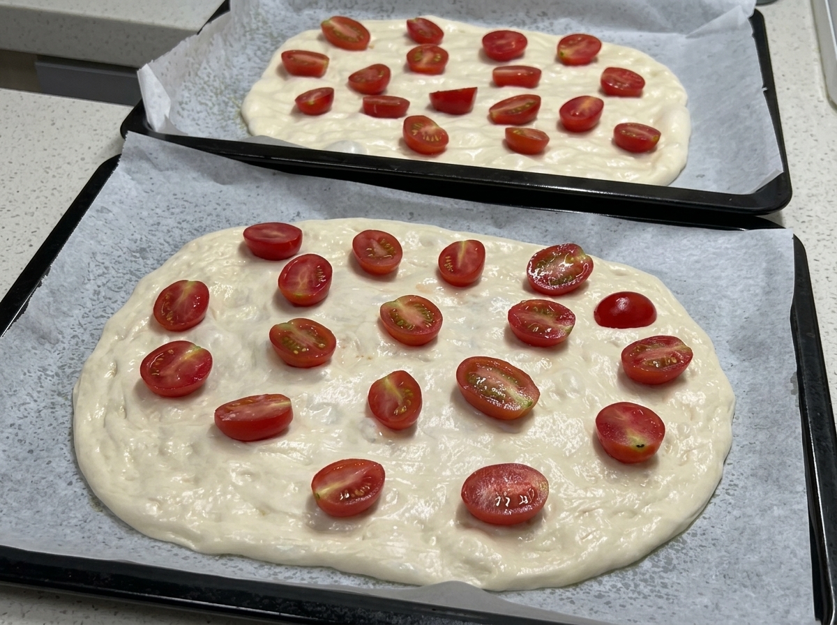 Se muestran dos bandejas de horno con masa de pizza extendida y cubiertas con numerosos tomates cherry partidos por la mitad, listas para sazonar.