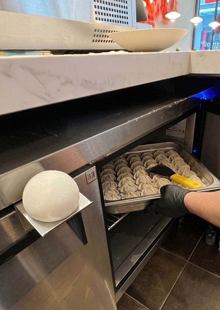 Una mano con guante saca una bandeja de acero inoxidable llena de porciones redondas y blancas de masa de pizza de una unidad de refrigeración profesional.
