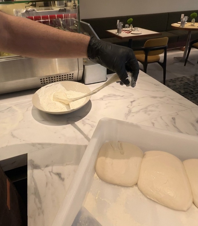Una mano con guante levanta una pieza elástica de masa de pizza fermentada de un recipiente con harina blanca.