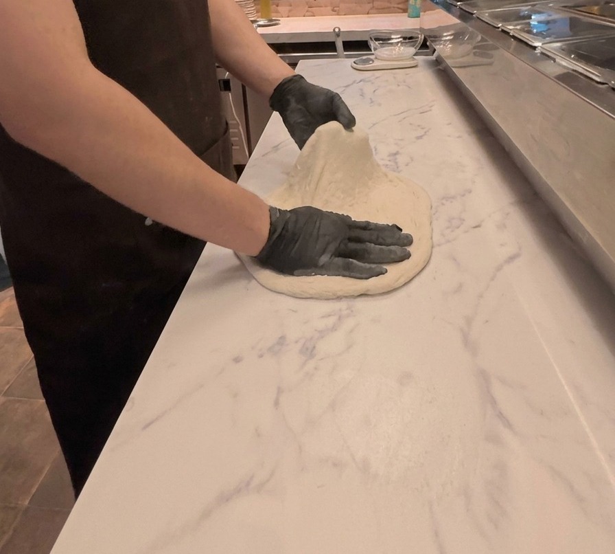 Un cocinero estira y aplana con ambas manos la masa de pizza sobre una encimera blanca con efecto mármol.