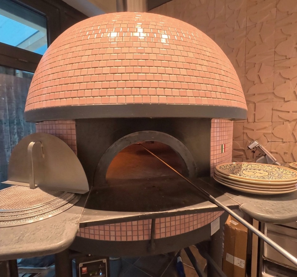 El exterior de un horno de pizza abovedado con azulejos rosas, con una pizza visible horneándose en su interior cerca de las brasas resplandecientes.