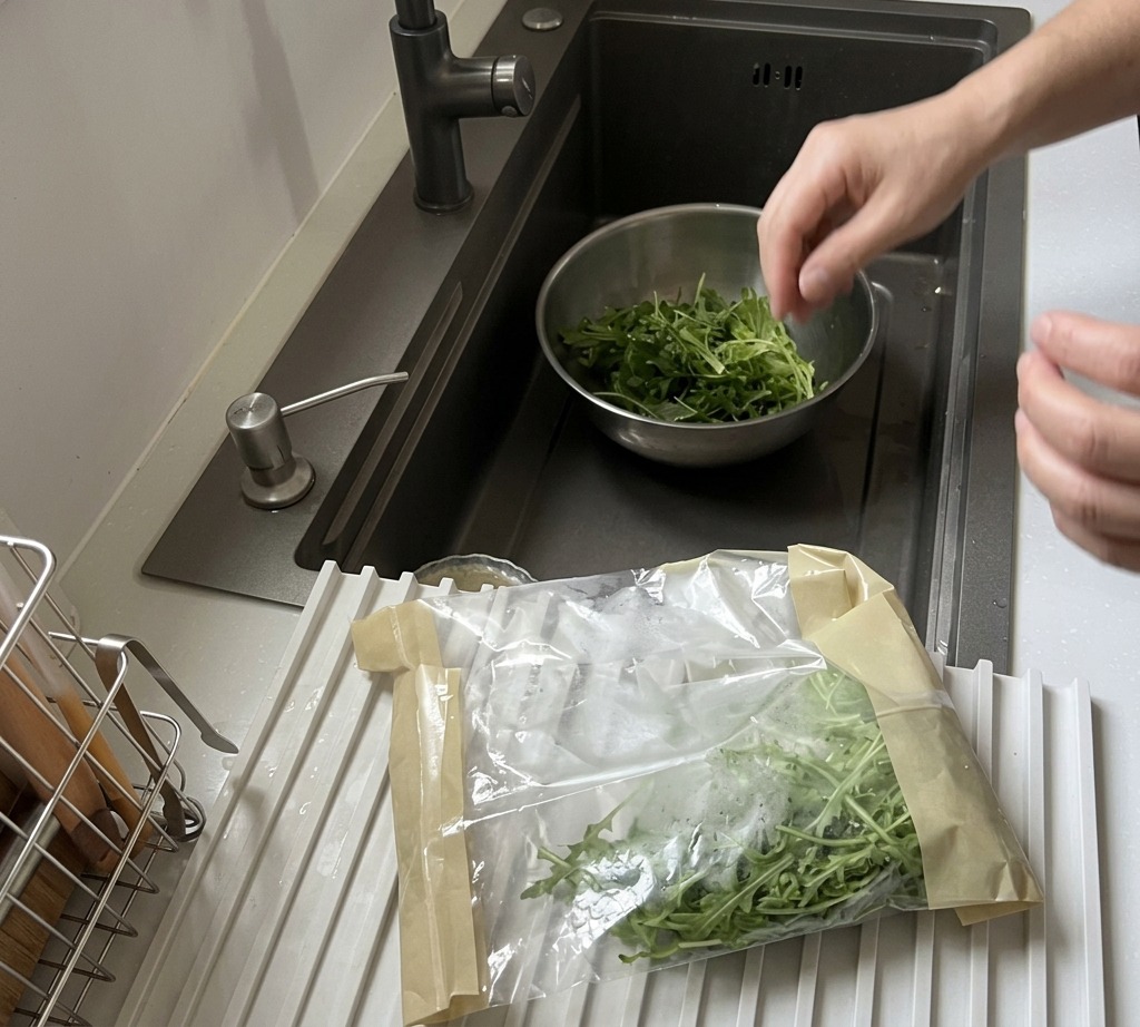 Hojas frescas de rúcula siendo lavadas en un tazón de acero inoxidable bajo el grifo de la cocina.