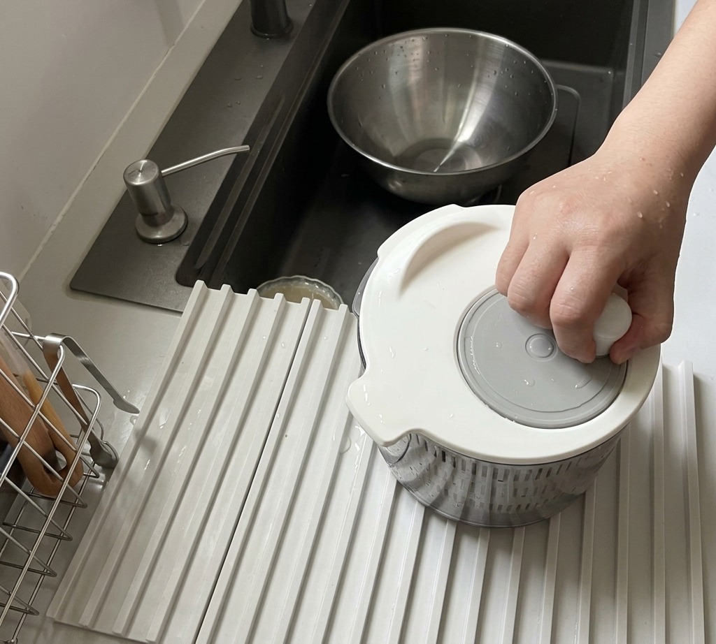 Una persona usando un centrifugador de ensalada manual blanco para secar hojas de rúcula frescas.