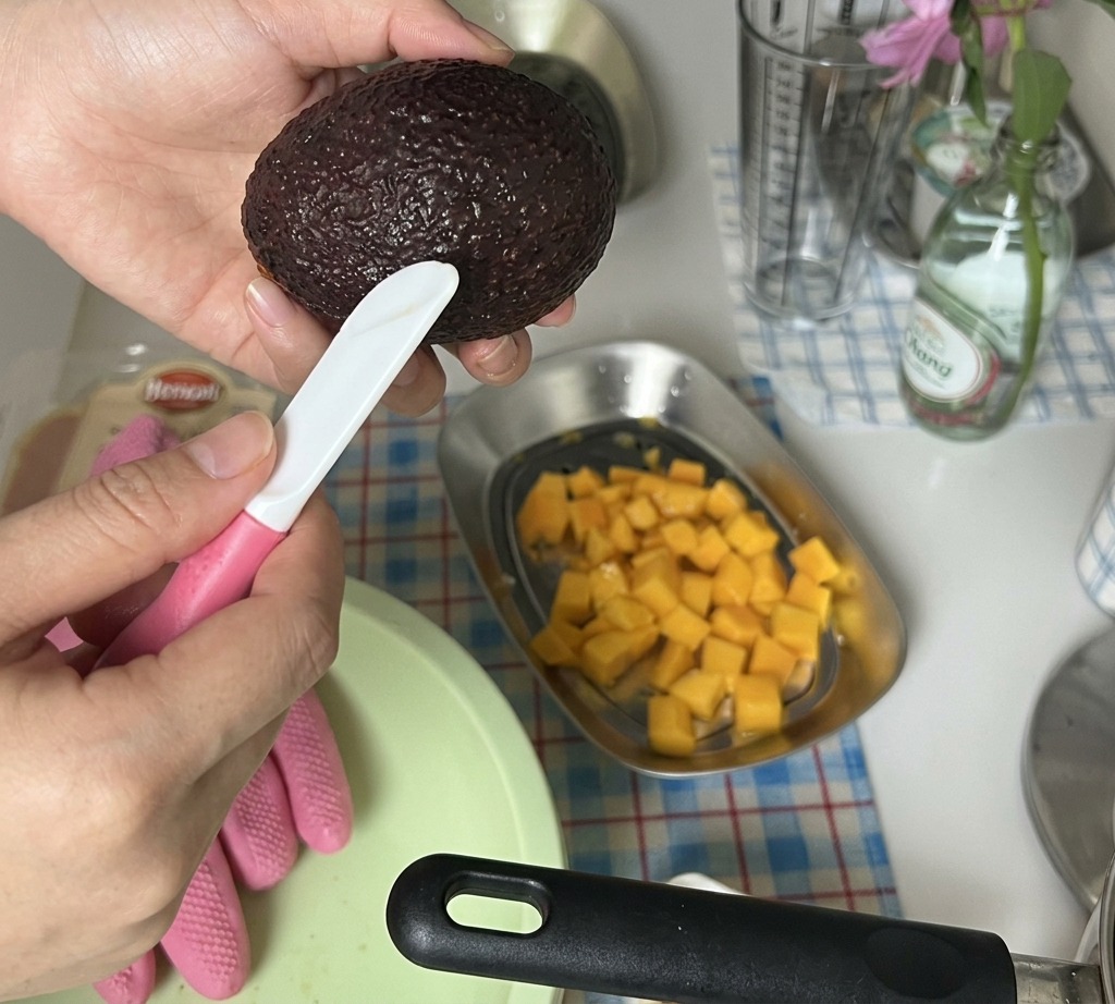 Manos cortando la mitad de un aguacate pelado en cubos pequeños sobre una bandeja con mango picado.