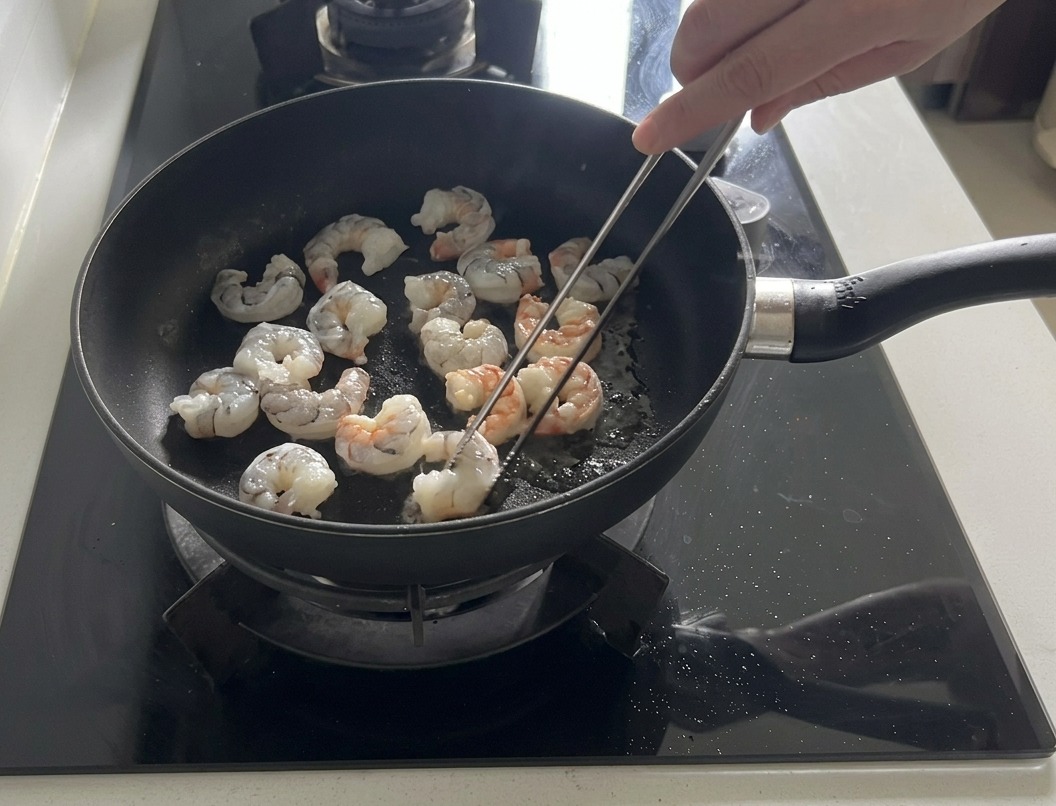 Camarones frescos siendo salteados en una sartén oscura con aceite de oliva, algunos volviéndose rosados y opacos.