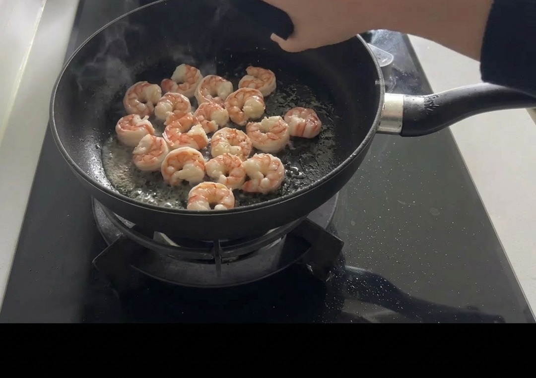 Camarones rosados salteados en una sartén negra siendo sazonados con sal y pimienta a mano.