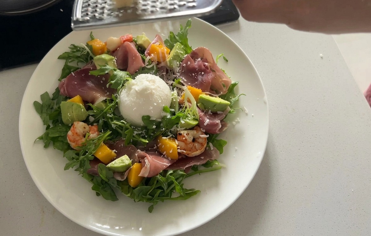 Una ensalada de rúcula emplatada coronada con una gran pieza de queso burrata blanco y finas láminas de prosciutto curado.