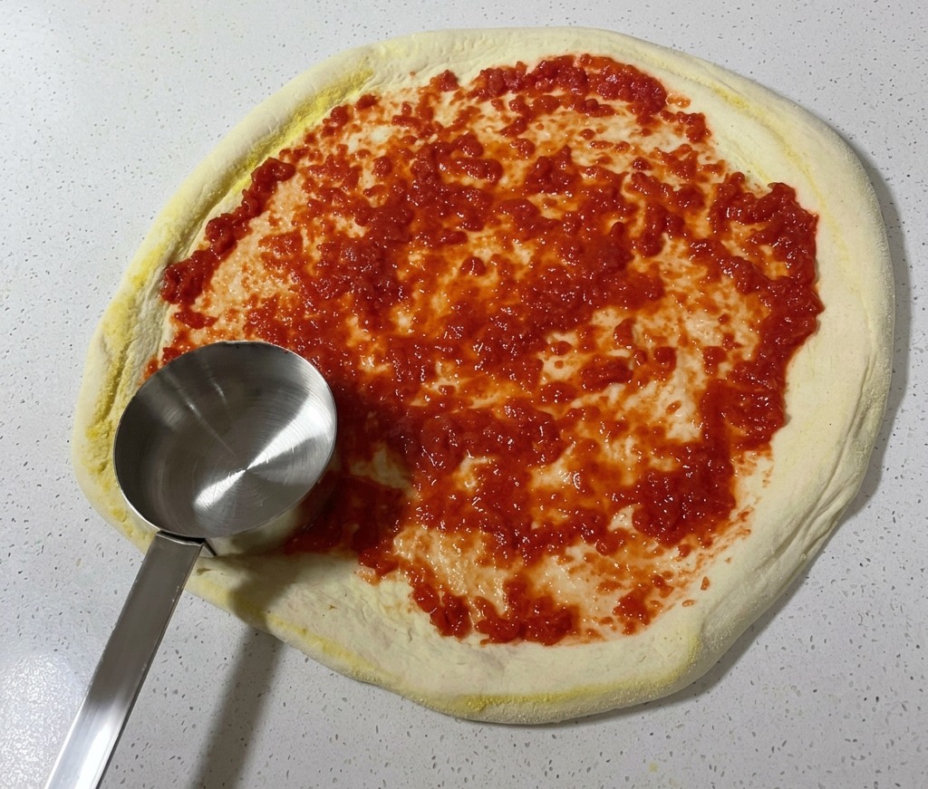 Un cucharón de metal extendiendo salsa de tomate de color rojo vibrante en forma circular sobre la superficie de la masa de pizza cruda.