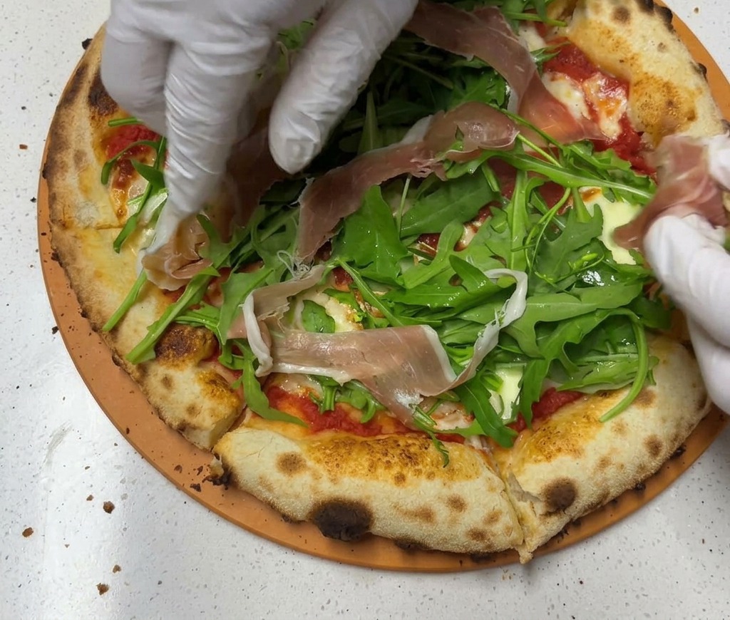 Manos con guantes colocando finas lonchas de jamón de Parma rosado sobre una cama de rúcula verde en una masa de pizza horneada.