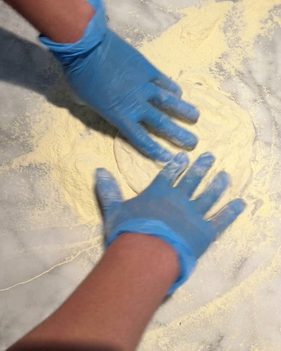 Manos con guantes azules presionando una bola de masa de pizza sobre una base abundante de sémola en una encimera de mármol.
