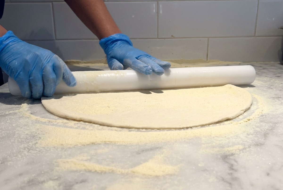 Un rodillo blanco estirando una base fina de masa de pizza sobre una mesa de mármol espolvoreada con harina.
