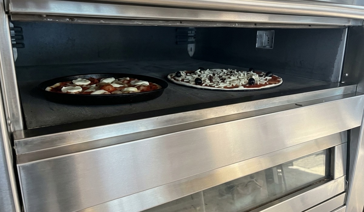 Dos pizzas horneándose sobre una superficie de piedra dentro de un horno de pisos profesional de acero inoxidable.
