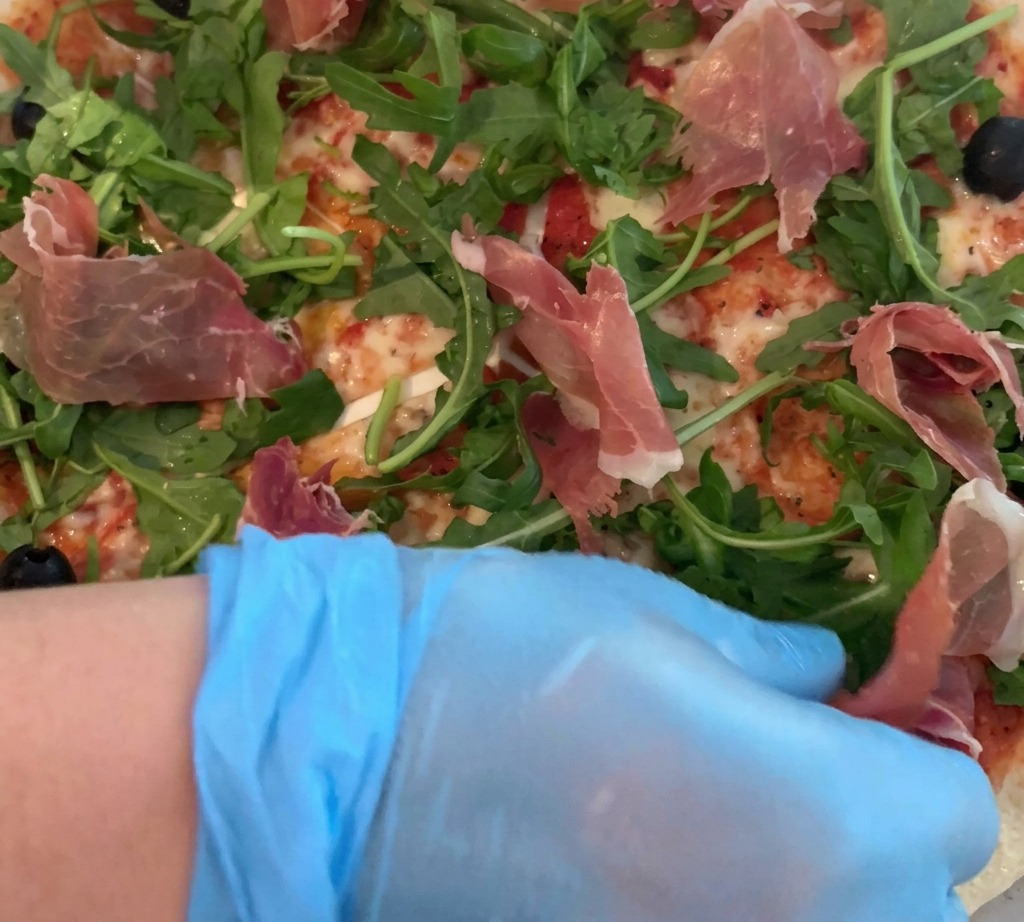 Una mano con guante colocando cuidadosamente finas lonchas de prosciutto sobre una pizza con rúcula fresca.