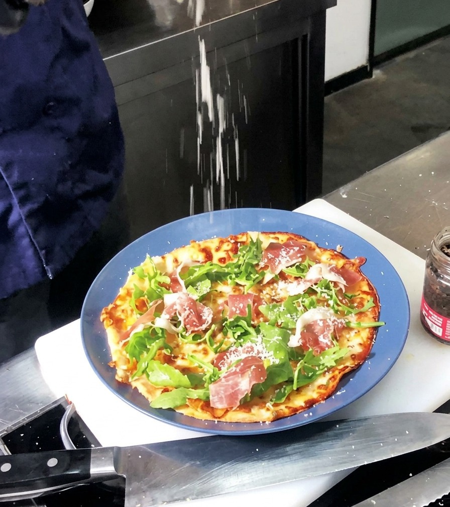 Polvo fino de queso parmesano blanco espolvoreado sobre una pizza horneada con rúcula verde y lonchas de jamón de color rojo.