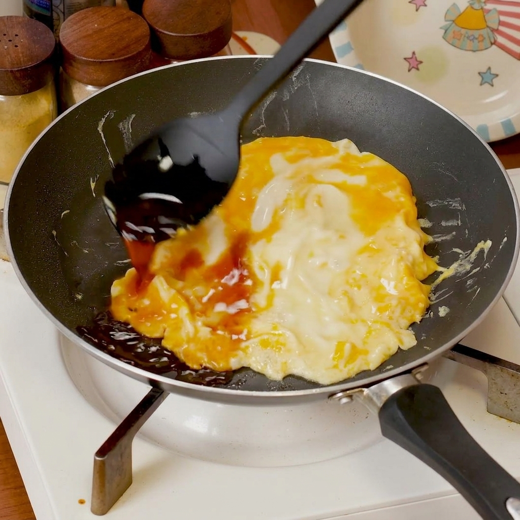 Salsa salada siendo vertida sobre huevos revueltos suaves cocinándose en una sartén.