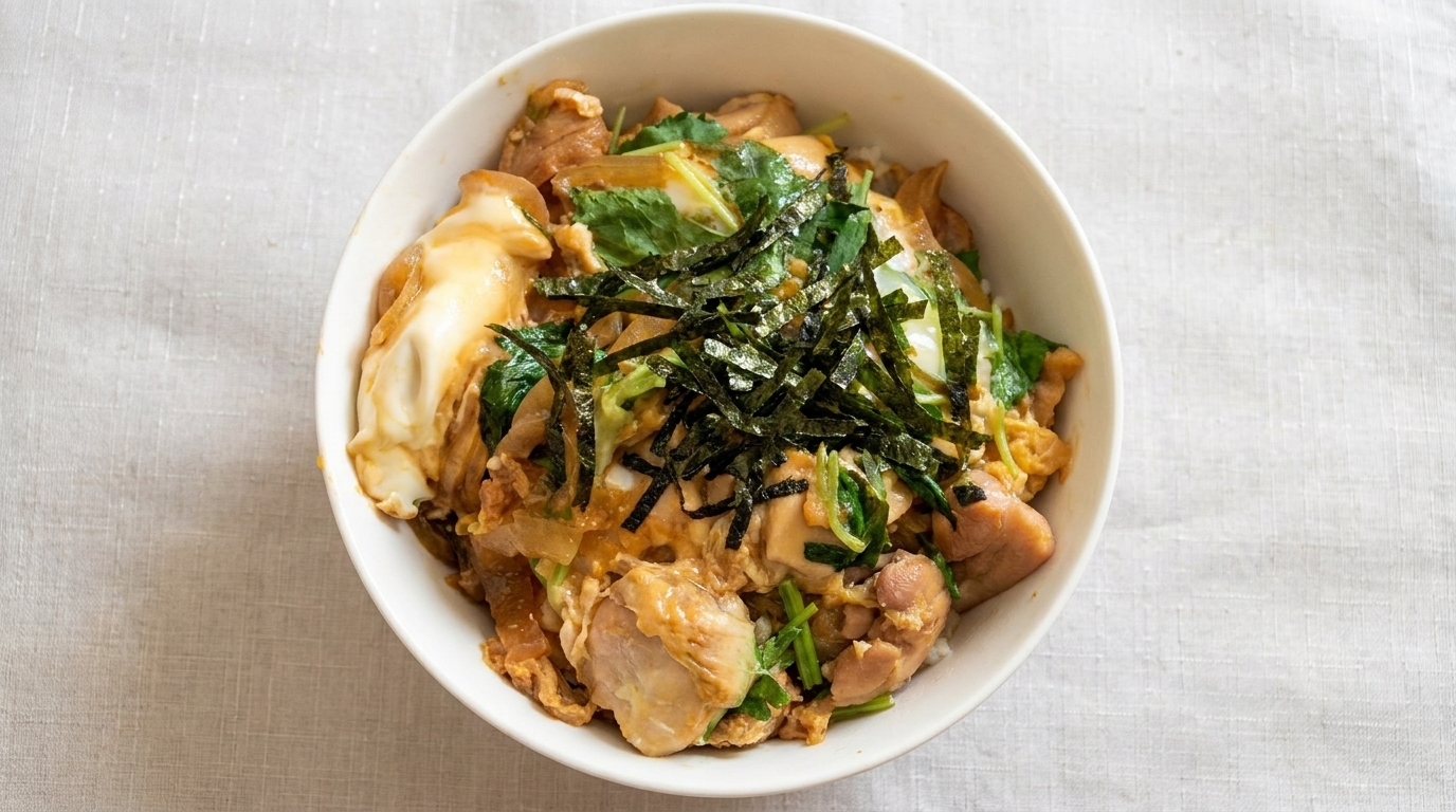 Un cuenco clásico de Oyakodon con pollo tierno y huevos cocinados a punto, decorado con mitsuba fresco y alga nori rallada.