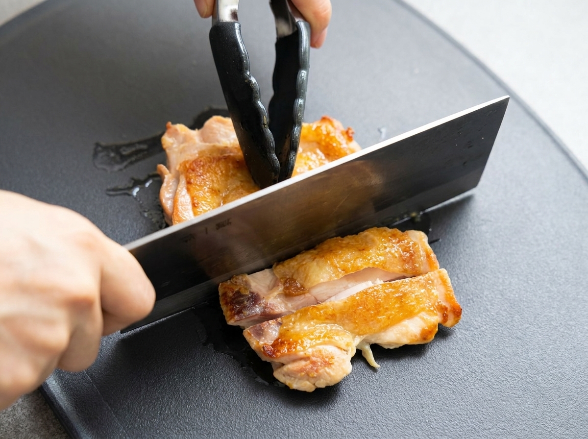 Muslo de pollo dorado siendo cortado en trozos sobre una tabla de cortar oscura.