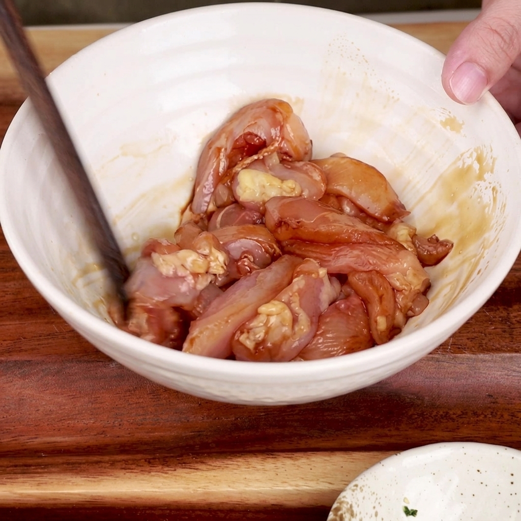 Unos palillos de madera mezclando trozos de pollo marinado en un bol blanco.