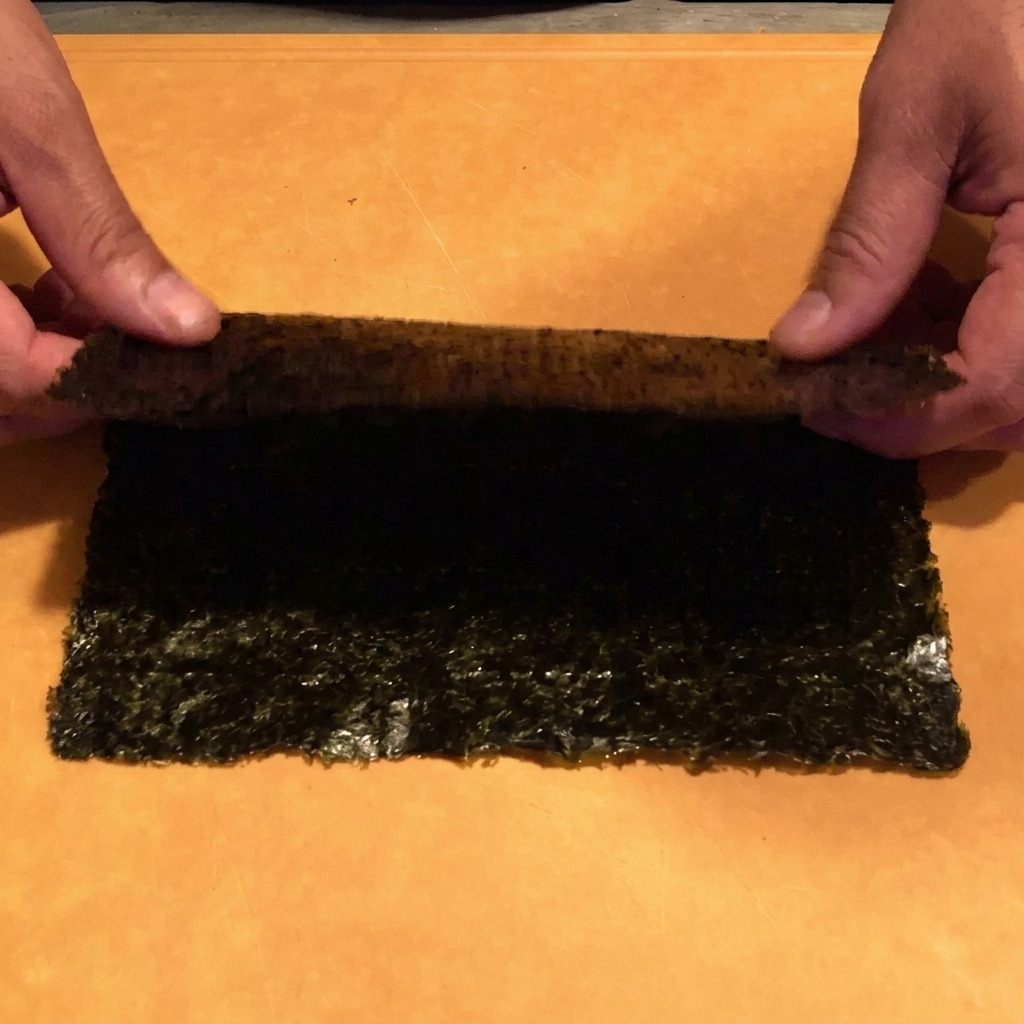 Manos cortando una hoja de alga nori en tiras finas con unas tijeras de cocina sobre un cuenco.