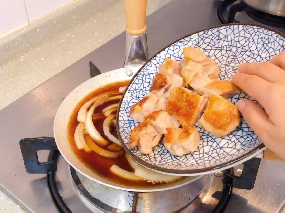 Rodajas de pollo crujiente a la sartén añadiéndose a una sartén con cebollas a fuego lento y salsa a base de soja.