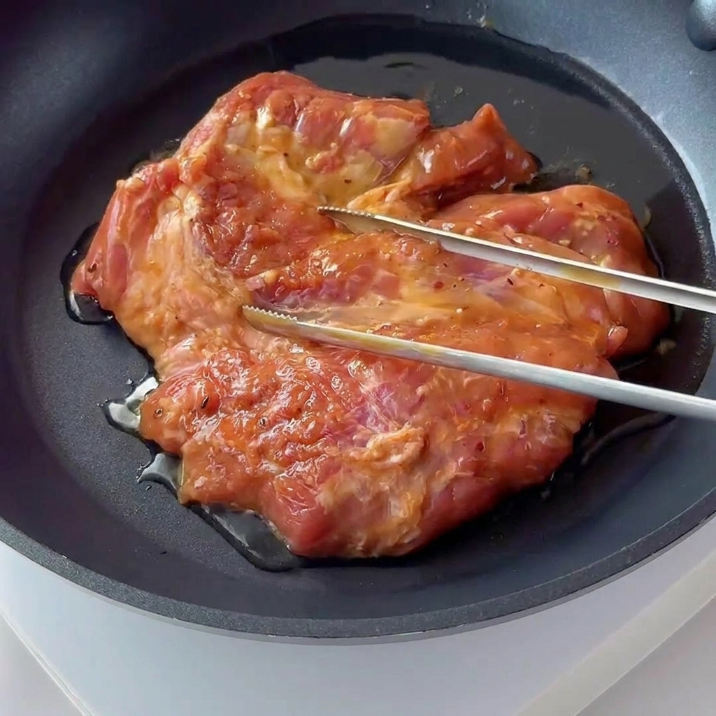 Una chuleta de cerdo marinada dorándose y chisporroteando en una sartén oscura antiadherente.
