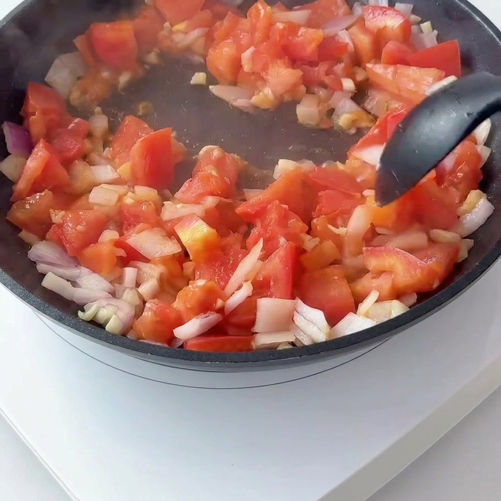 Tomates en cubitos y cebolla roja salteándose en una sartén mientras se mezcla la pasta de tomate.