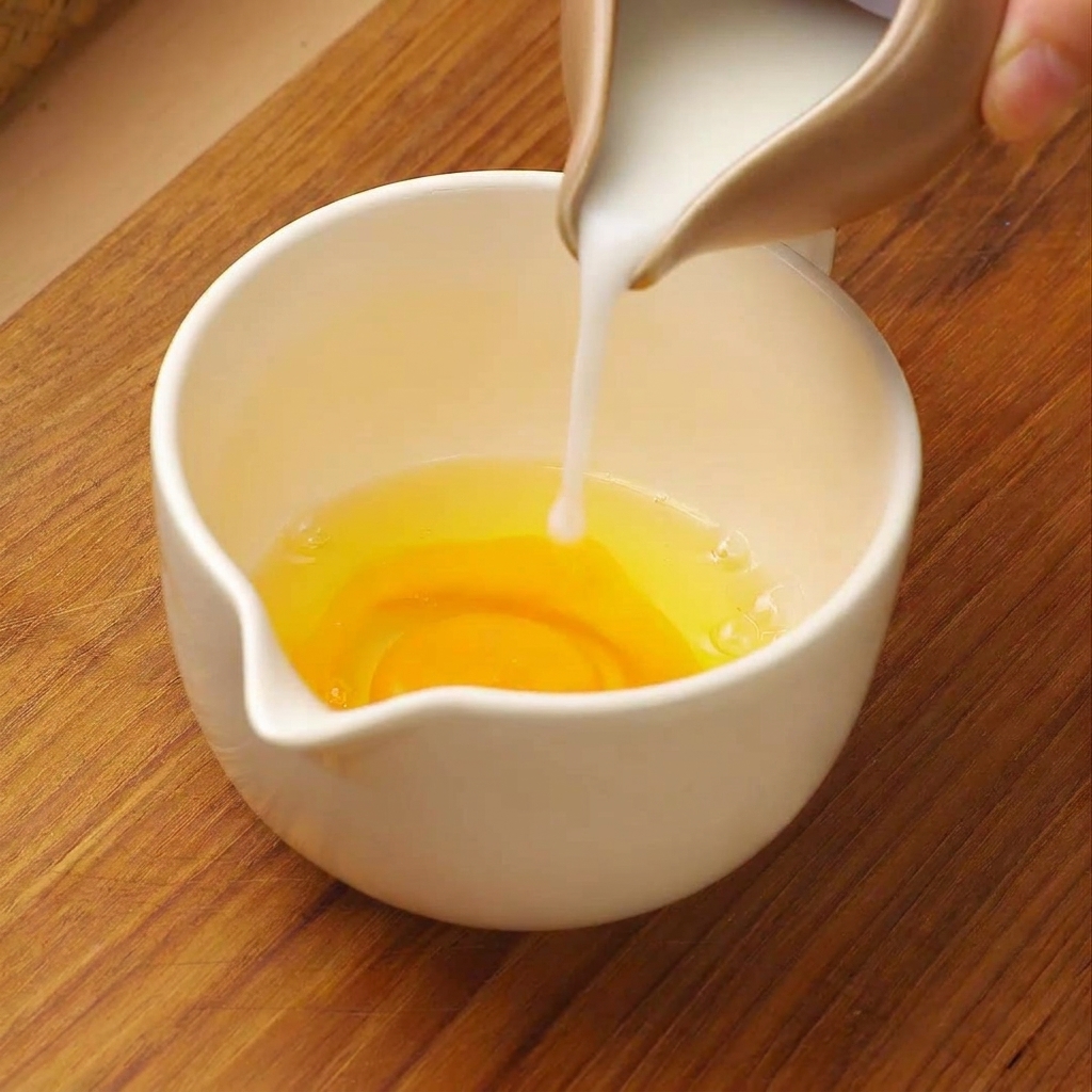 Leche blanca vertiéndose desde una jarra pequeña en una taza medidora blanca que contiene huevos crudos.