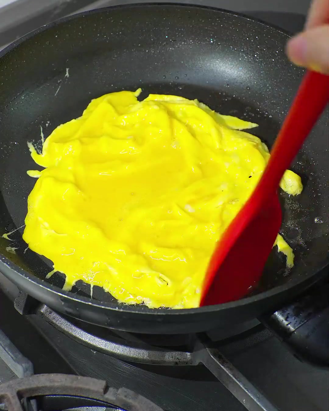 Una espátula roja doblando un huevo revuelto de color amarillo brillante y parcialmente cocido en una sartén negra.