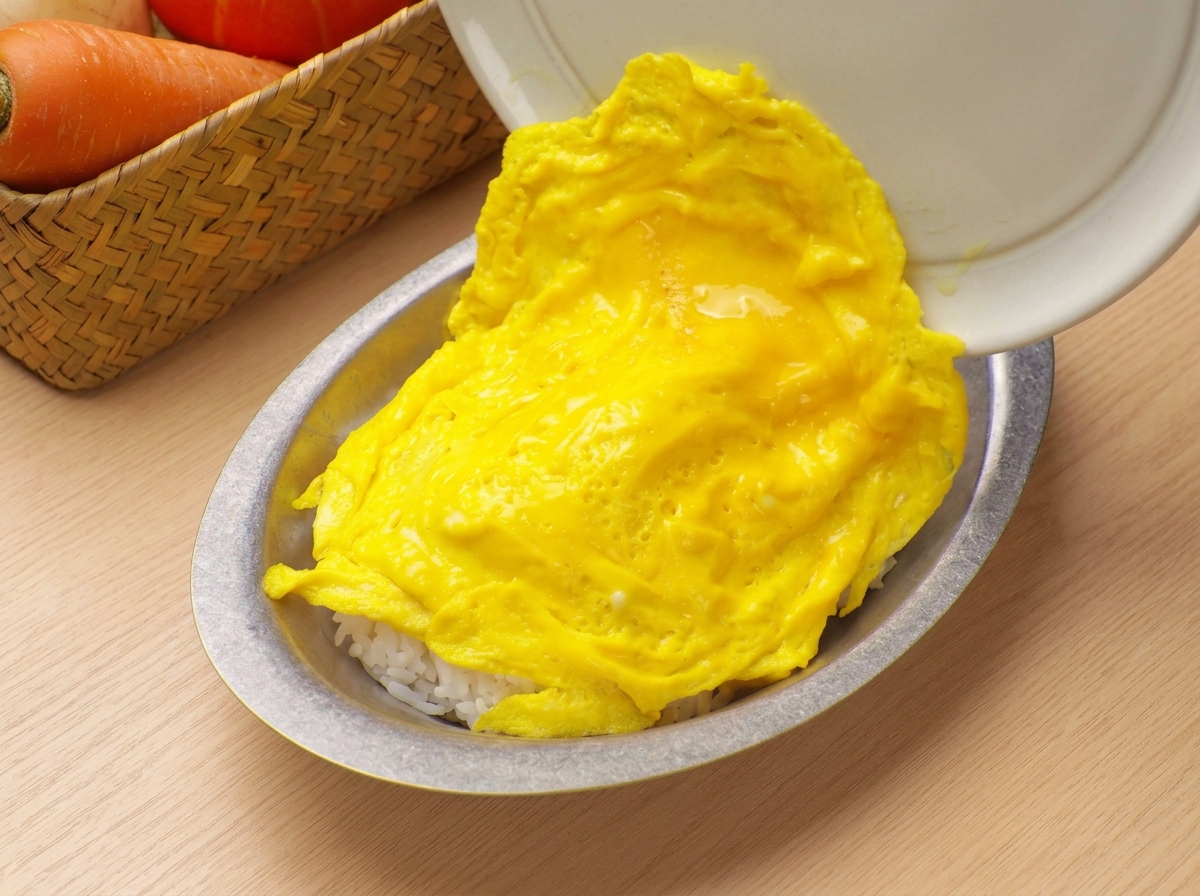 Huevos revueltos suaves y esponjosos deslizándose desde una sartén hacia un bol de servicio lleno de arroz blanco.