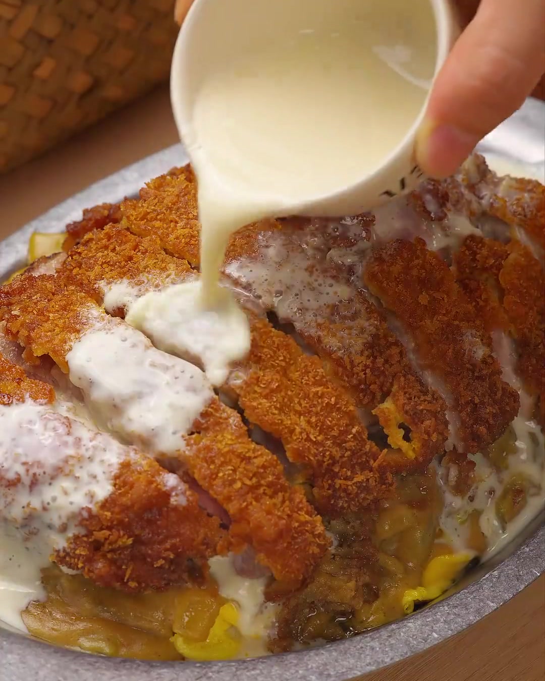 Un primer plano de una cremosa salsa blanca de queso y coco vertiéndose sobre chuletas de cerdo en rodajas doradas, adornada con perejil verde fresco.