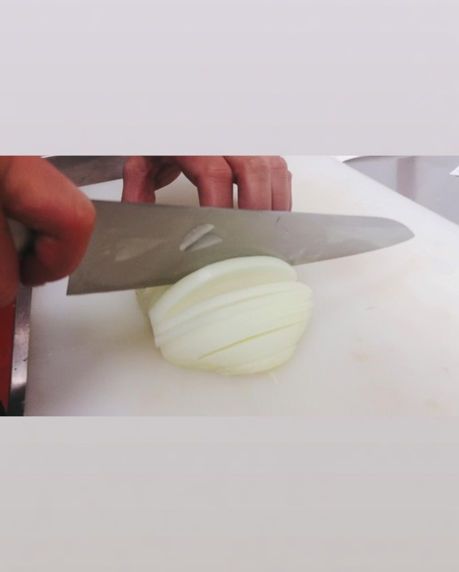 Cortando una cebolla blanca cruda con un cuchillo grande sobre una tabla de cortar blanca.