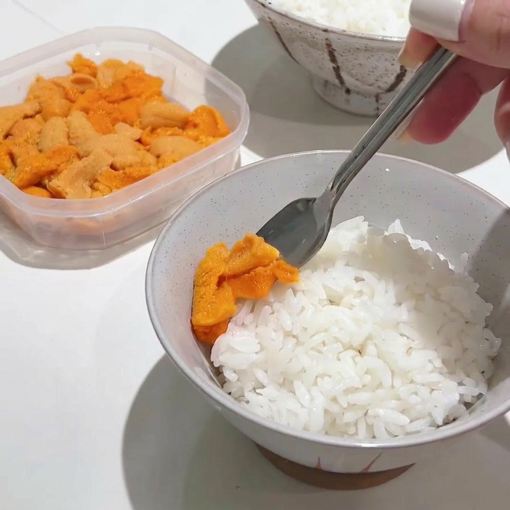 Una cuchara de metal colocando suavemente una porción generosa de erizo de mar naranja brillante sobre un cuenco de arroz blanco caliente.