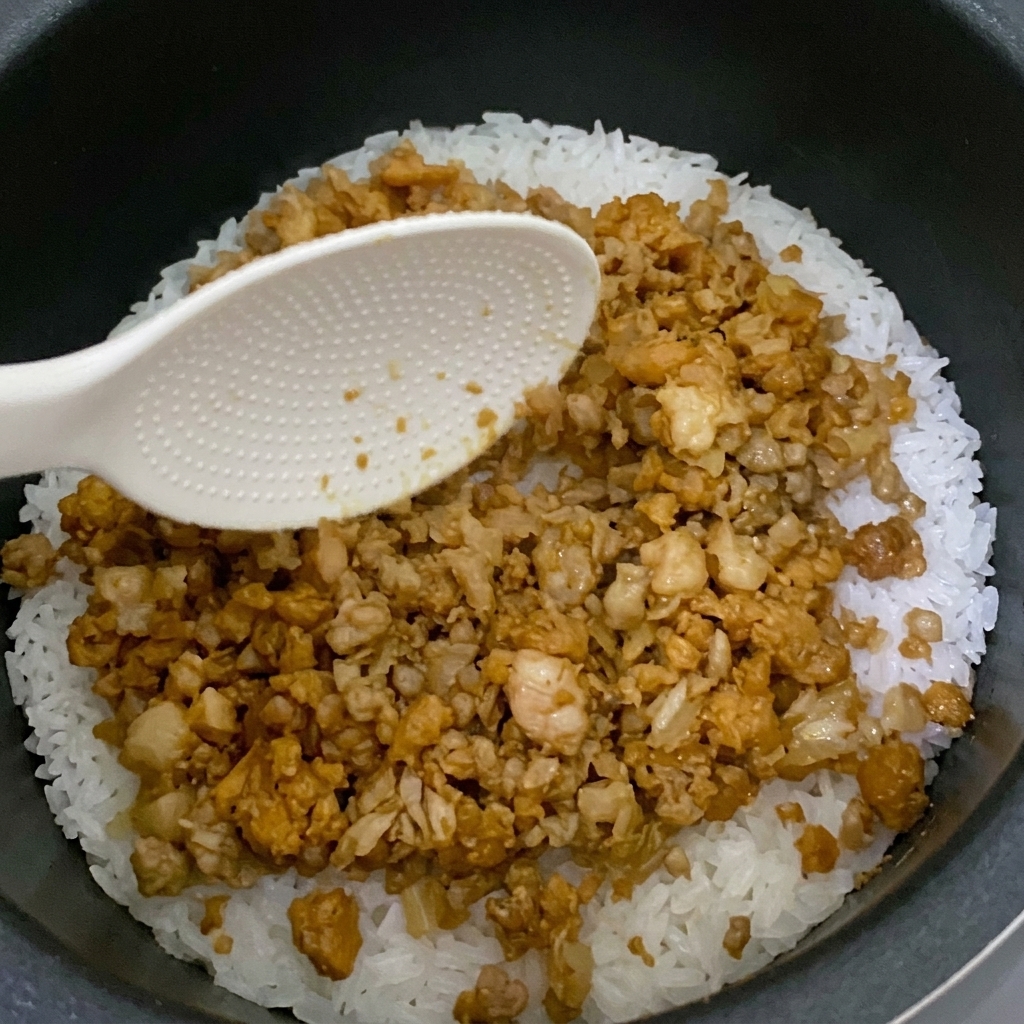 La mezcla salteada terminada extendiéndose sobre el arroz en una olla arrocera.