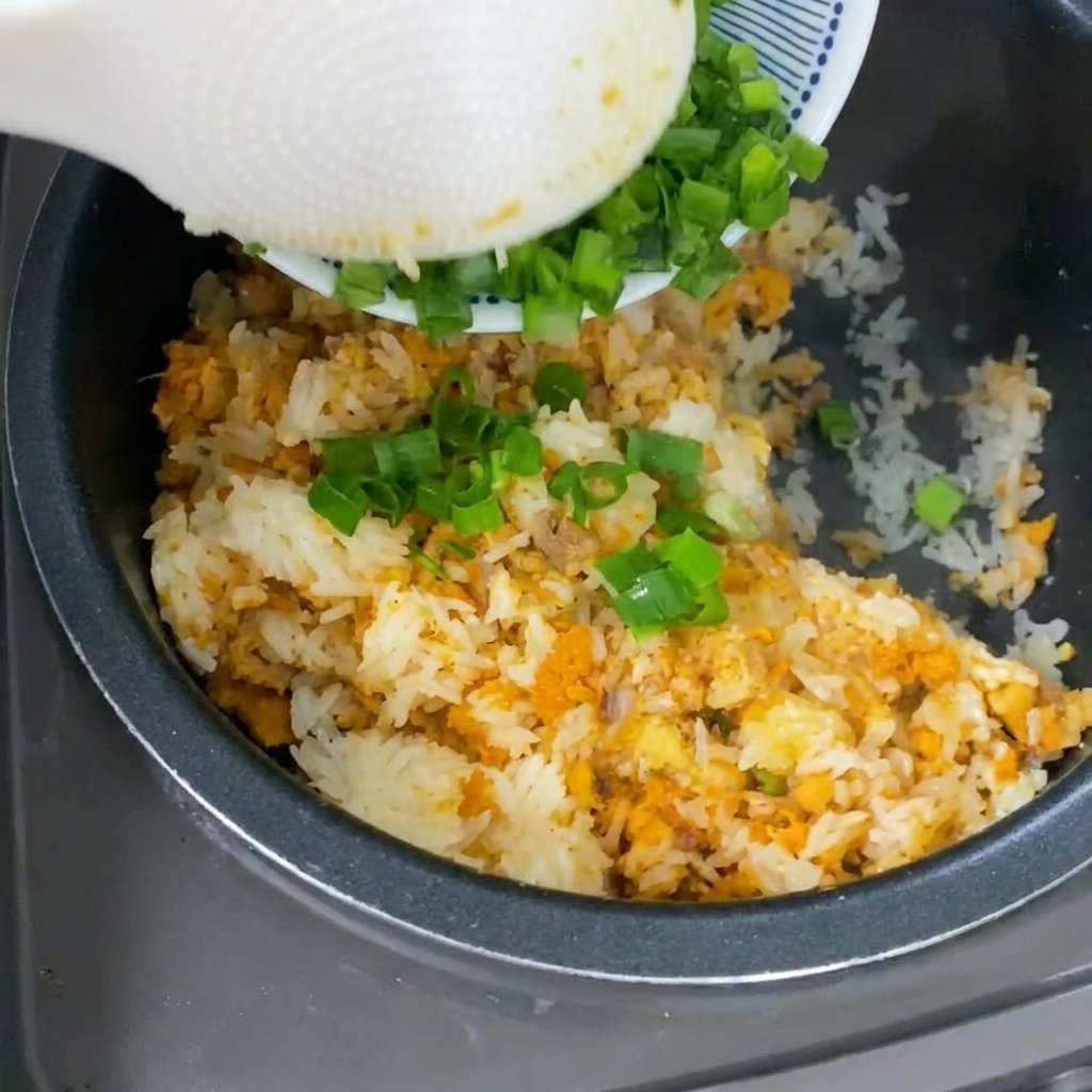 Una cuchara blanca vertiendo cebolletas verdes recién picadas sobre el arroz cocido de tono amarillo dentro de una olla arrocera.