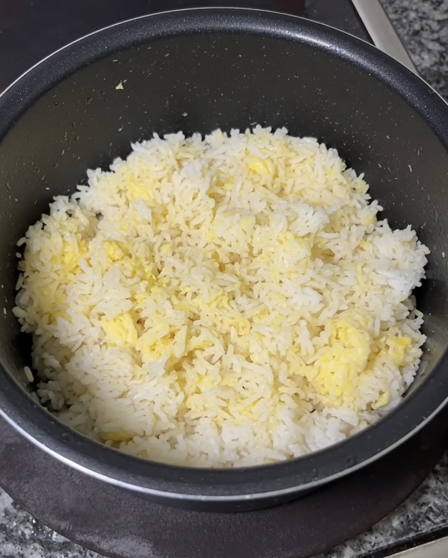 Una mano usando una cuchara para mezclar a fondo yemas de huevo crudas en un tazón de arroz blanco frío hasta que quede uniformemente amarillo.