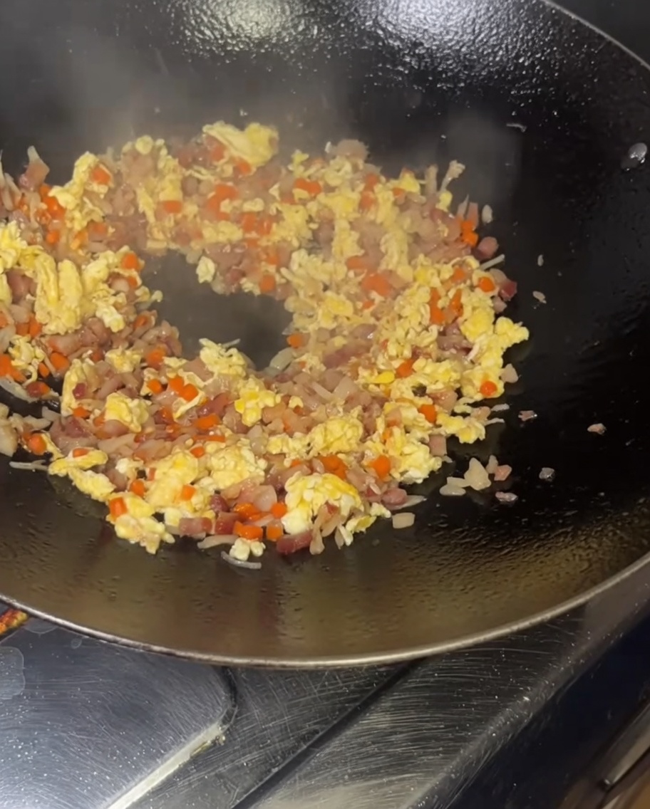 Vaciando un tazón de granos de arroz amarillos y cubiertos de huevo en un wok humeante y muy caliente, lleno de ingredientes salteados.