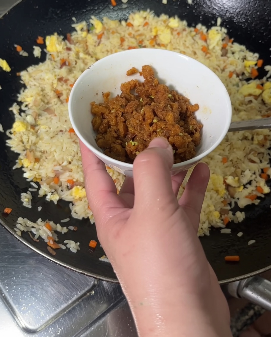 Una mano vertiendo huevas anaranjadas brillantes de erizo de mar ya cocidas desde un pequeño tazón hacia un wok lleno de arroz frito.