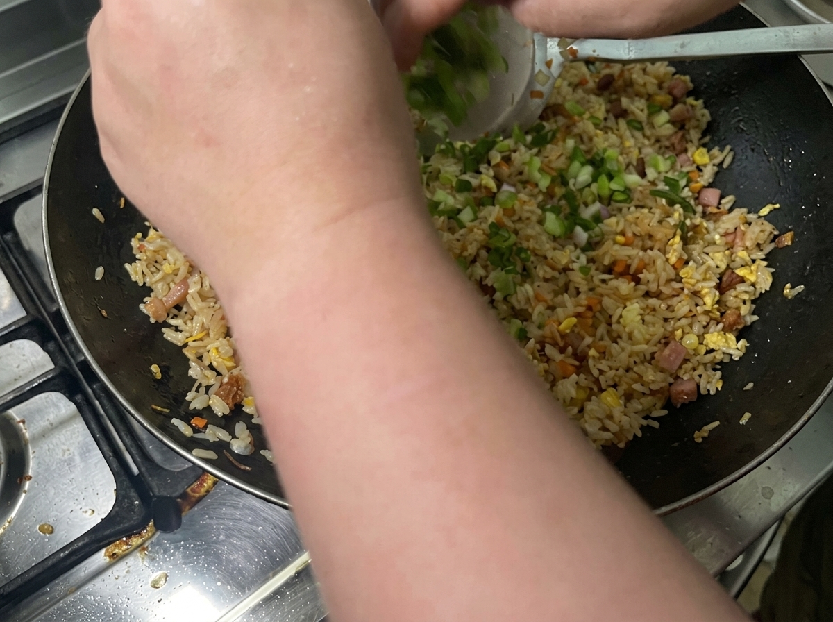 Cebolletas picadas siendo arrojadas a un wok lleno de arroz frito sazonado.
