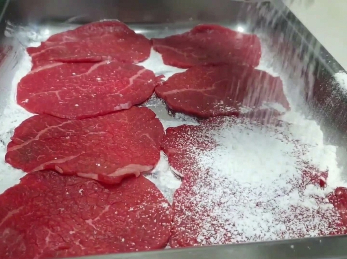 Mano espolvoreando harina blanca sobre rebanadas de ternera en una bandeja.