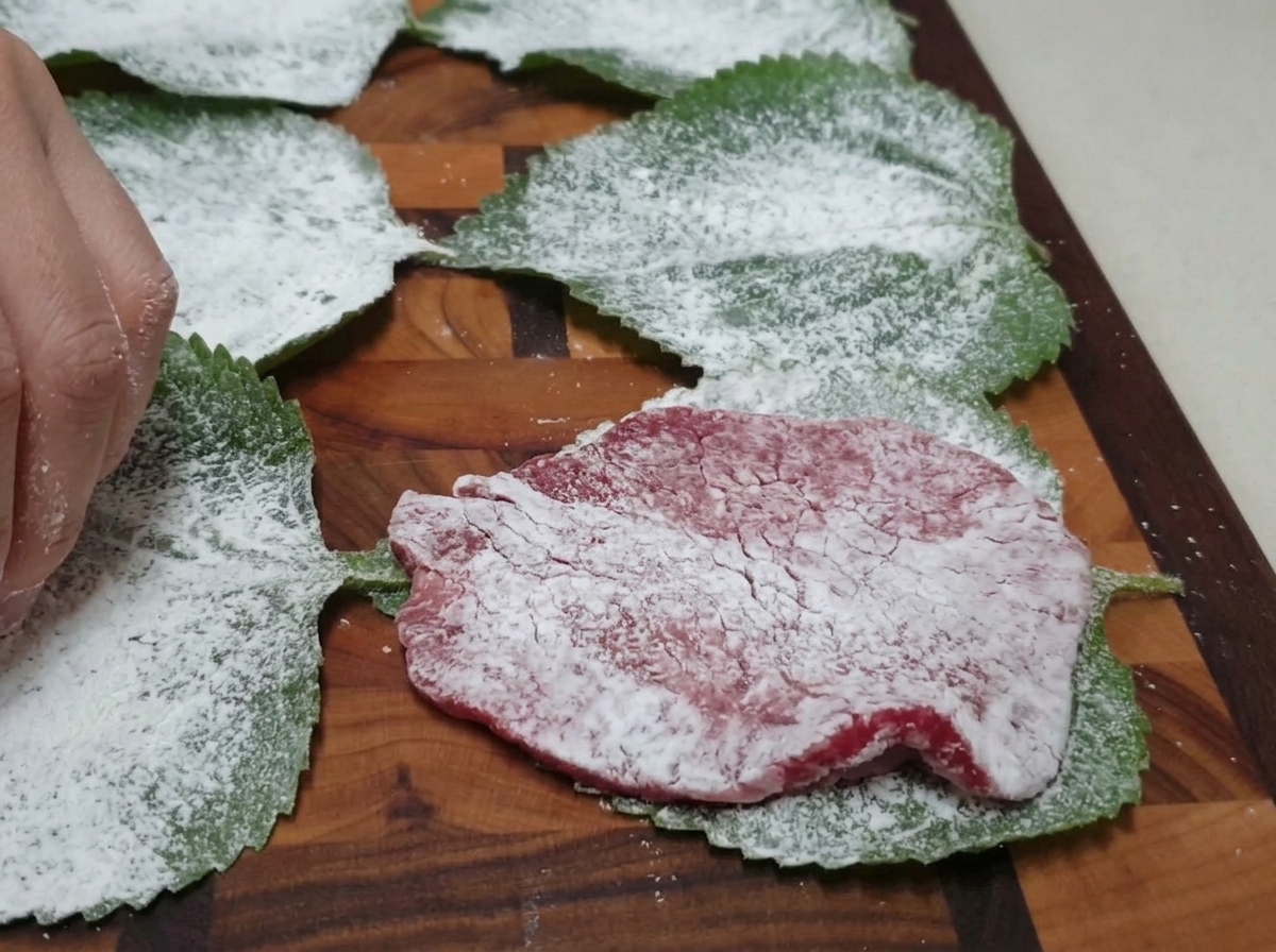 Una rebanada de ternera enharinada colocada sobre una hoja de perilla en una tabla de cortar de madera.