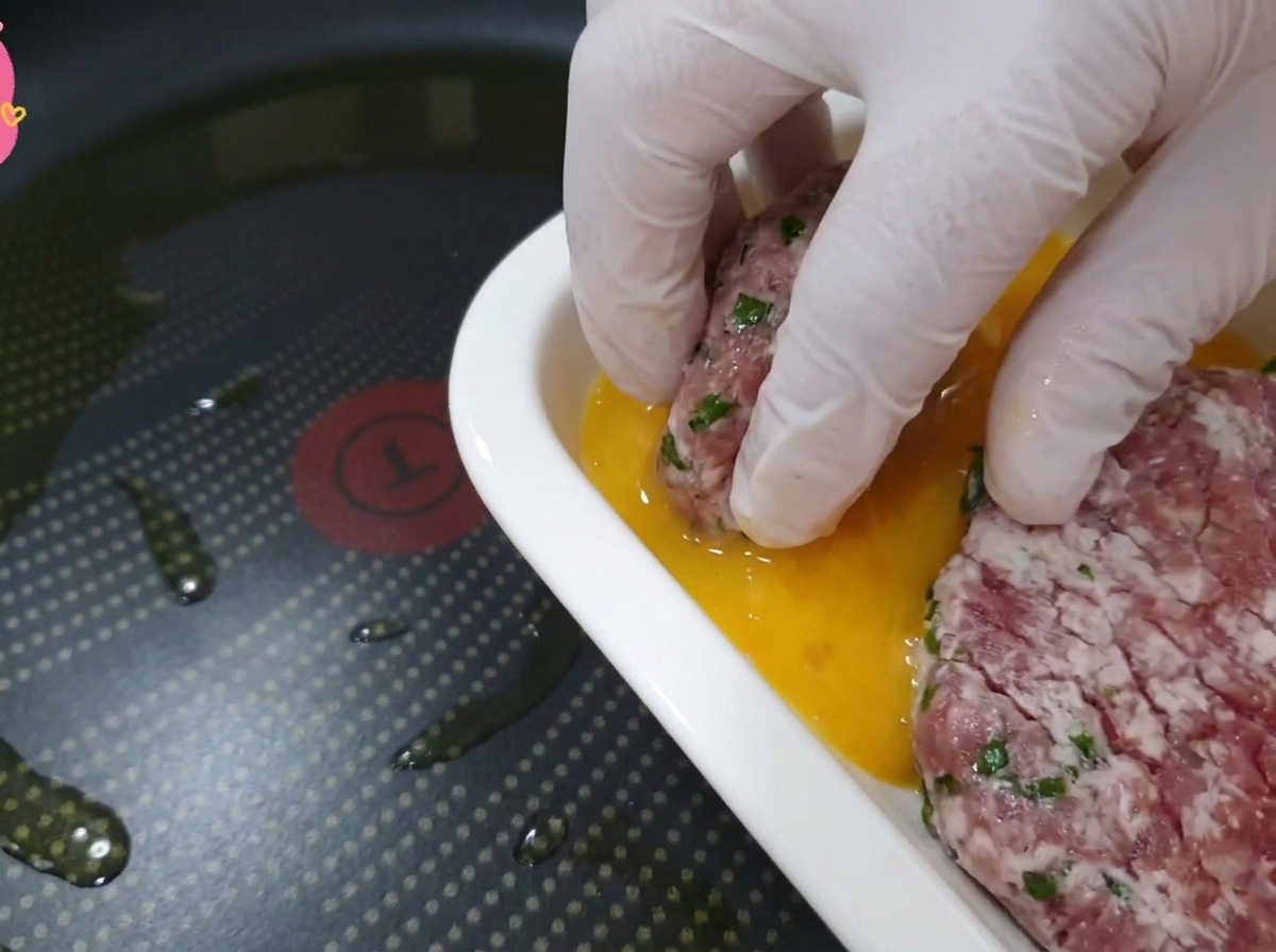 Mano con guante transparente sumergiendo la carne con la hoja de perilla en un plato con huevo batido dorado.